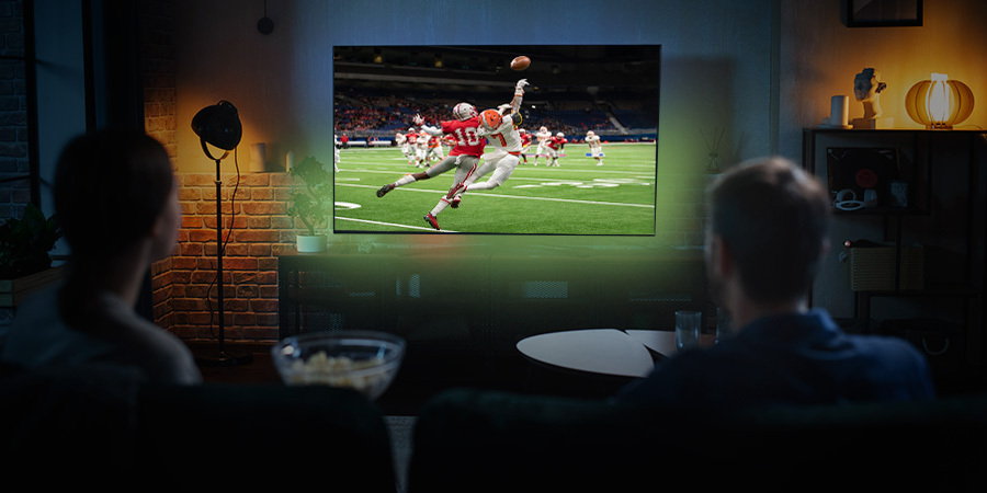 couple-watching-a-football-game-at-home A couple sitting on a couch in a cozy living room, watching a football game on a large television, with a bowl of popcorn on the table in front of them.