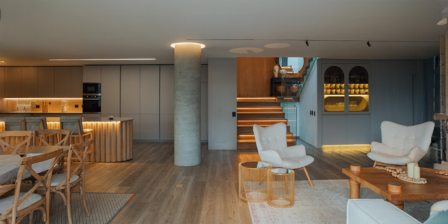 modern-house-with-lighting-design-using-white-led-strips Warm ligModern living and dining area with warm wooden floors and minimalist decor. LED strip lights accentuate a staircase, a ceiling column, and cabinets, creating a cozy ambiance. The space includes light-colored armchairs, a wooden dining table, and built-in shelving with soft lighting.