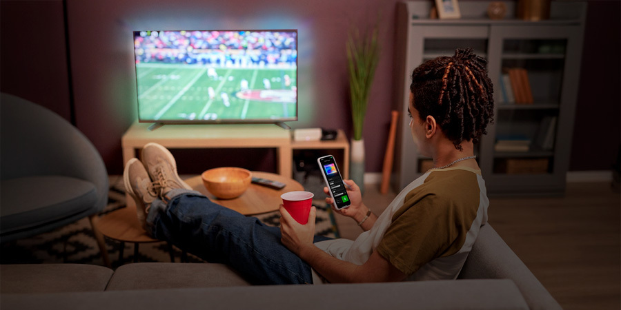watching-football-game-on-tv-while-controling-the lights-on-phone A young man relaxing on a couch, watching a football game on TV while holding a smartphone displaying the Leona Smart App
